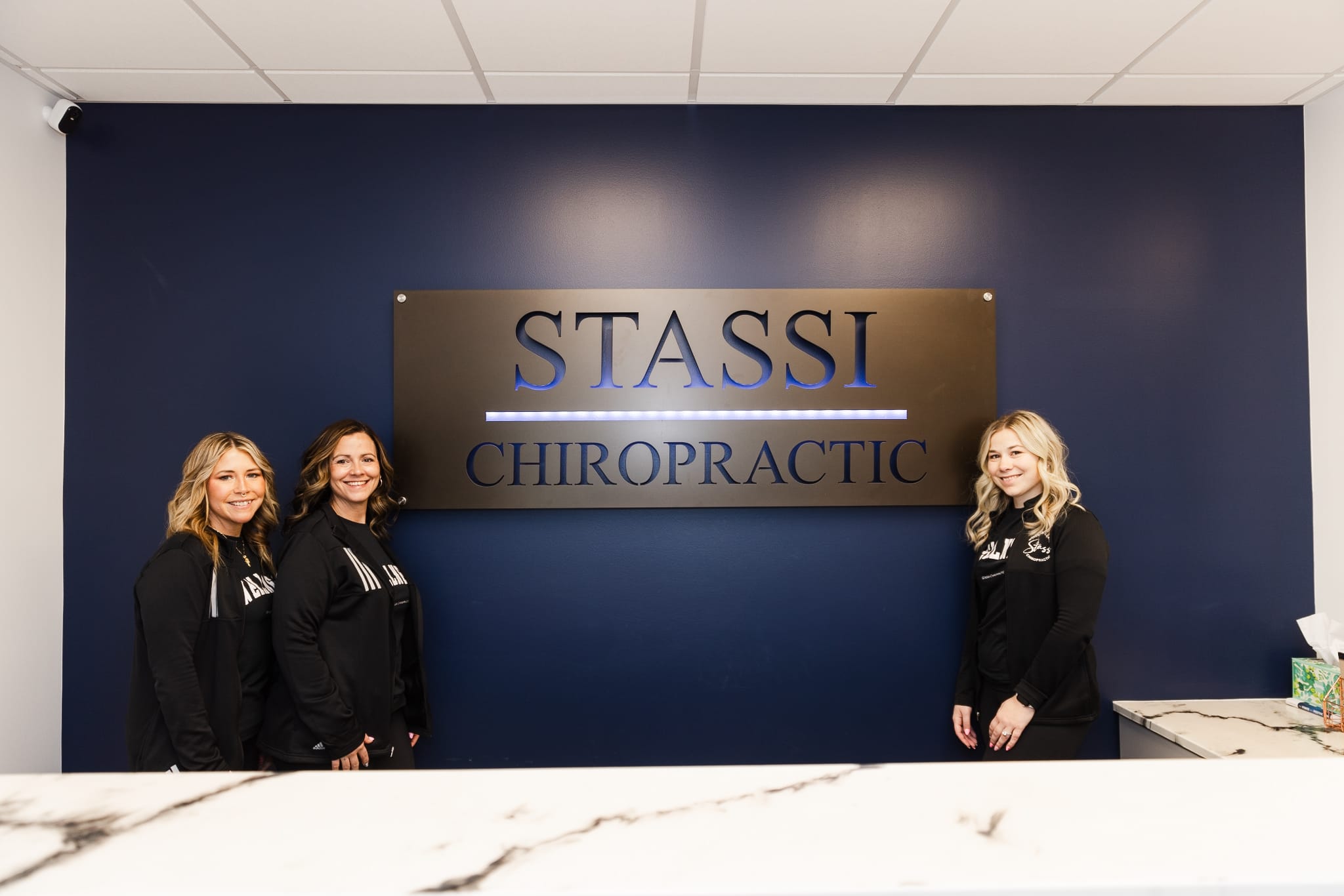 Three smiling team members, including one man and three women, in black Stassi Chiropractic branded apparel.